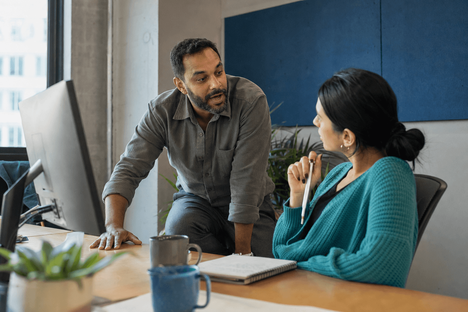 Two coworkers talking in front of a blurred monitor in a bright coworking space, she wearing a turquoise cardigan holding a pen and him explaining something with his hand resting on the desk.