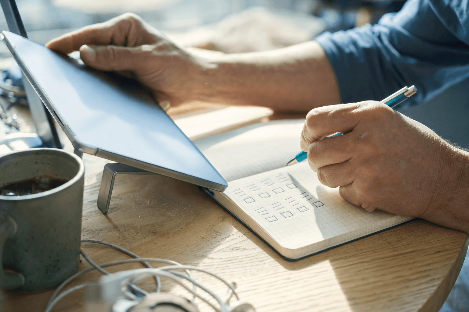 Close-up of a professional's hands holding a pen over a checkered notebook, next to a blurred tablet on a stand, with natural light from the window.